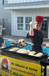 Poolside hibachi chef in a tall red hat and sunglasses theatrically sprinkling seasoning over rice on a flat-top griddle at a backyard outdoor catering setup.
