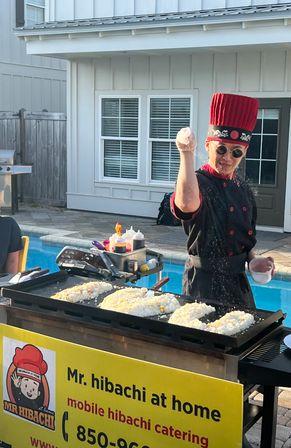Poolside hibachi chef in a tall red hat and sunglasses theatrically sprinkling seasoning over rice on a flat-top griddle at a backyard outdoor catering setup.