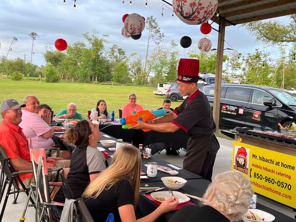 Mobile hibachi chef in a red hat entertaining seated guests under a pavilion at an outdoor backyard-style party, aiming a colorful water gun while diners laugh amid hanging paper lanterns and picnic tables