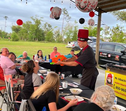 Mobile hibachi chef in a red hat entertaining seated guests under a pavilion at an outdoor backyard-style party, aiming a colorful water gun while diners laugh amid hanging paper lanterns and picnic tables