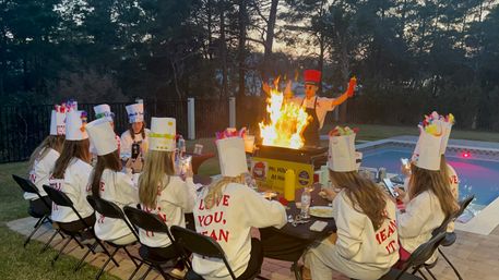 Poolside backyard cooking party at dusk: a chef ignites a dramatic flaming grill while guests in decorated paper chef hats and matching sweatshirts sit around a table filming the spectacle.