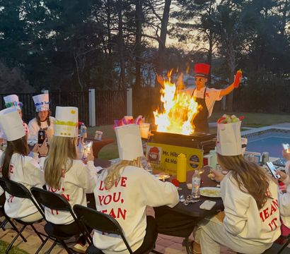 Poolside backyard cooking party at dusk: a chef ignites a dramatic flaming grill while guests in decorated paper chef hats and matching sweatshirts sit around a table filming the spectacle.