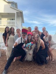 Group of friends posing outdoors in front of a white coastal beach house under a blue sky, some wearing novelty tall red chef hats and apron and holding a colorful toy water gun — playful vacation group photo.