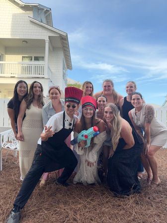 Group of friends posing outdoors in front of a white coastal beach house under a blue sky, some wearing novelty tall red chef hats and apron and holding a colorful toy water gun — playful vacation group photo.