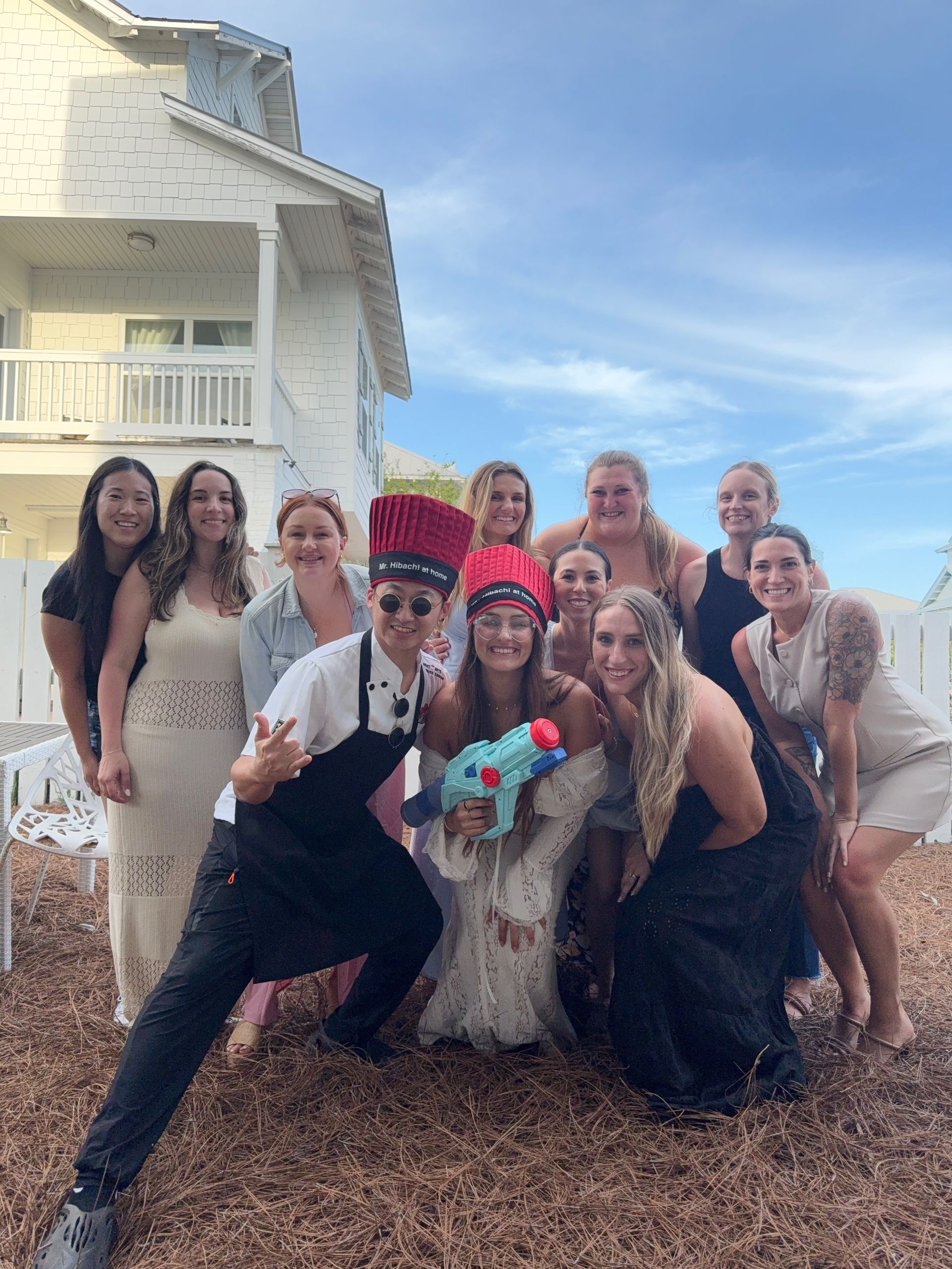 Group of friends posing outdoors in front of a white coastal beach house under a blue sky, some wearing novelty tall red chef hats and apron and holding a colorful toy water gun — playful vacation group photo.