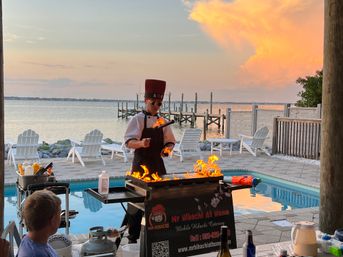 Poolside hibachi chef in a tall hat cooking over dramatic flames at sunset by a calm bay and dock, with a pool, Adirondack chairs, and onlookers nearby.