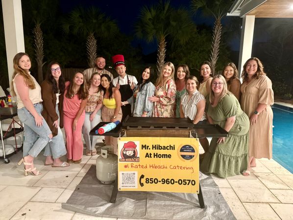 Cheerful group of adults posing around a portable hibachi grill and a chef in a red hat at a backyard pool party at night, palm trees in the background and a yellow catering sign on the grill.