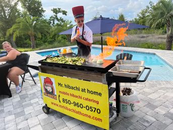 Poolside mobile hibachi chef in a tall red hat and sunglasses theatrically ignites a flaming flat-top grill while cooking vegetables on a catering cart beside a backyard pool and palm trees.