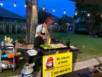 Evening backyard hibachi chef in a red tall hat flambéing vegetables and chicken on a mobile outdoor griddle under string lights