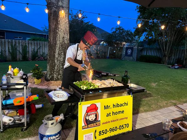 Evening backyard hibachi chef in a red tall hat flambéing vegetables and chicken on a mobile outdoor griddle under string lights