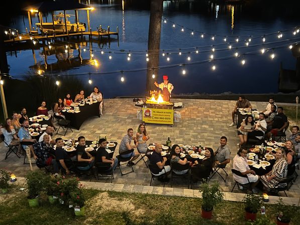 Evening waterfront patio dinner under string lights with a hibachi chef performing over a flaming grill, guests seated at long tables on a stone terrace with a dock and boat reflecting in the water