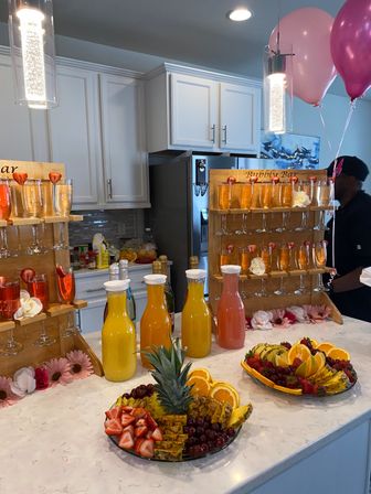 Home kitchen brunch mimosa bar on marble island with wooden flute racks, carafes of orange and pink juices, pineapple and strawberry fruit platters and pink balloons