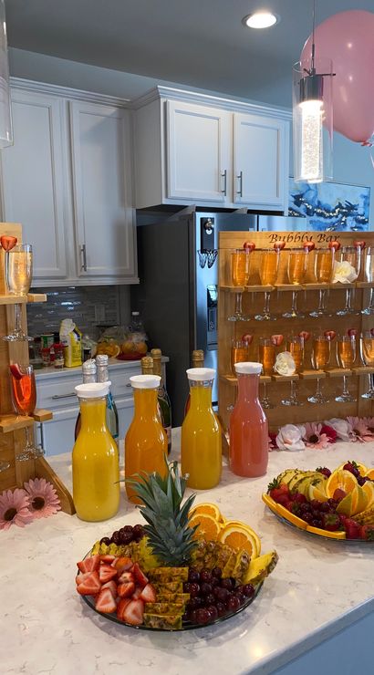 Home kitchen brunch mimosa bar on marble island with wooden flute racks, carafes of orange and pink juices, pineapple and strawberry fruit platters and pink balloons