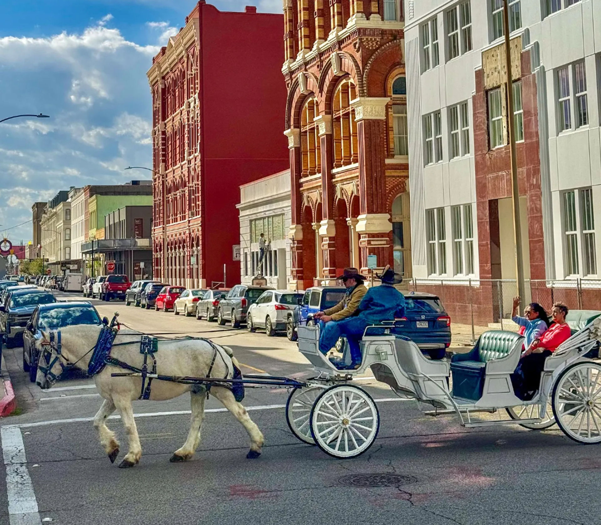White horse pulling a white sightseeing carriage with passengers past ornate red-brick historic downtown buildings on a sunny urban street