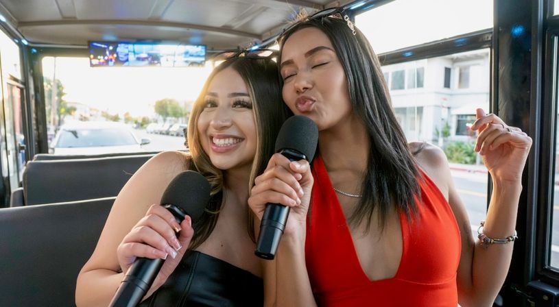Two friends duetting with microphones on a sunny party-bus karaoke ride, one in a red dress puckering and the other smiling, city street visible through the windows.