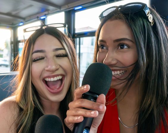 Two friends laughing and singing into microphones on a party-bus karaoke ride, close-up with sunglasses on their heads