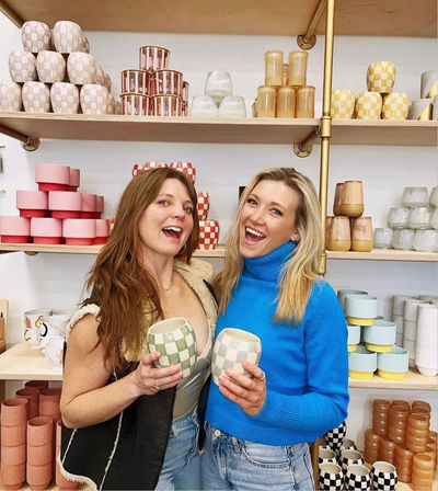 Two smiling women holding green-and-white checkered ceramic mugs in a bright boutique pottery shop with shelves of pastel and checkerboard cups and stacked pottery.