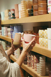 Shopper's hands with red nail polish comparing a matte pink tumbler and a speckled stoneware cup on wooden retail shelves filled with colorful ceramic mugs and vases.