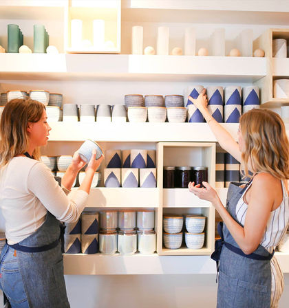 Two people arranging blue-and-white handmade ceramic bowls, jars and candles on white display shelves in a bright, minimalist home decor boutique