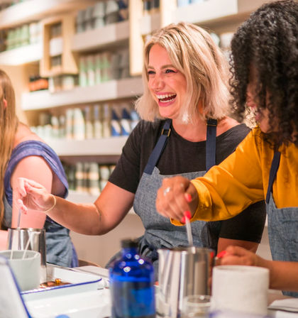 Smiling women in aprons stirring wax in metal pouring pots during a bright candle-making craft workshop