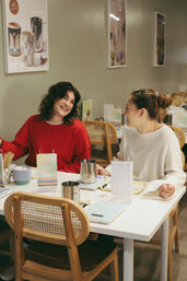 Two friends laughing over drinks at a cozy modern café table with wooden cane-back chairs, menus and stainless-steel pitchers