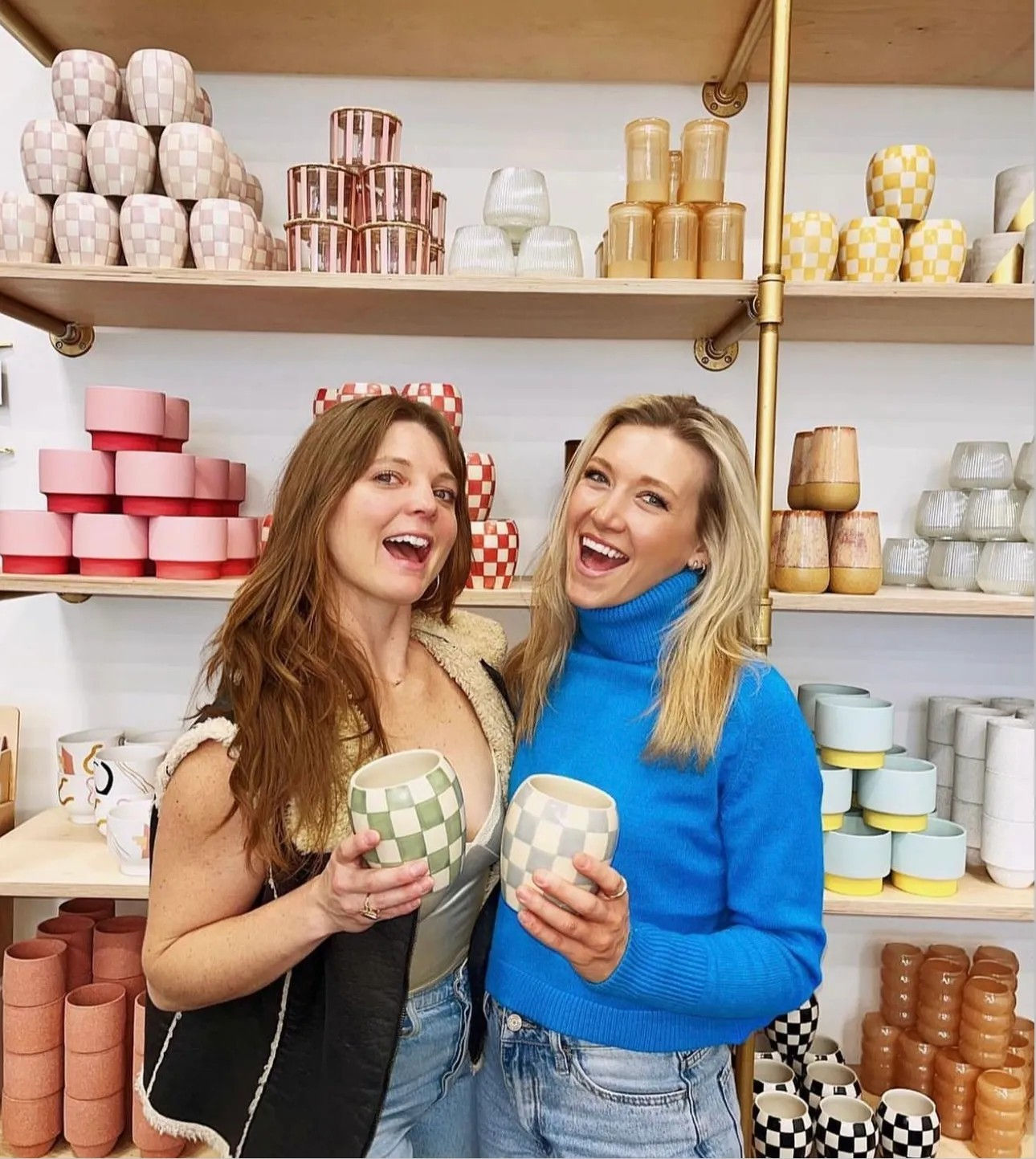 Two smiling women holding pastel checkered ceramic mugs in a bright boutique pottery shop with colorful stacked cups and vases on wooden shelves.