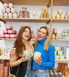 Two smiling women holding checkered ceramic mugs in a bright boutique pottery shop, posing in front of shelves stacked with pastel and patterned cups, planters, and colorful ceramics