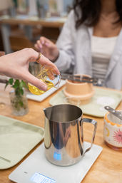 Hand pouring golden liquid from a small glass measuring cup into a stainless steel milk pitcher on a digital scale at a cafe table, blurred mug and tray in background.