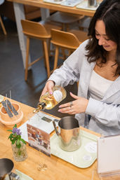 Person pouring pale fragrance oil into a glass beaker on a digital scale during a cozy candle-making workshop at a bright wood studio table, with a metal pouring pitcher, wick holders and a small vase of purple flowers.