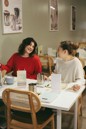 Two friends laughing across a white table in a cozy modern café with light-wood chairs, metal milk jugs, menus and tableware — casual coffee or brunch meet-up.