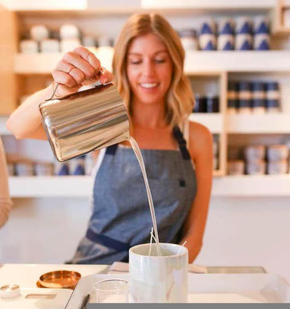 Smiling barista in an apron pouring steamed milk from a stainless-steel pitcher into a ceramic mug at a bright café counter