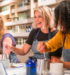 Smiling women in aprons stirring wax in metal tins during a lively artisan candle-making workshop at a bright craft studio