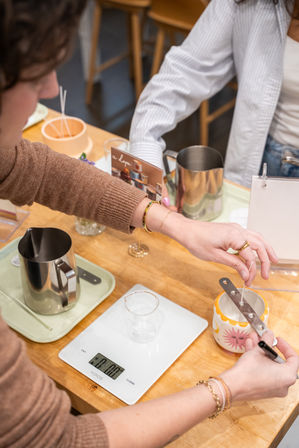 Participant hands placing a wick in a colorful ceramic candle jar with a sun motif using a metal wick holder at a candle-making workshop, with a digital kitchen scale, stainless steel pitcher and tools on a wooden table