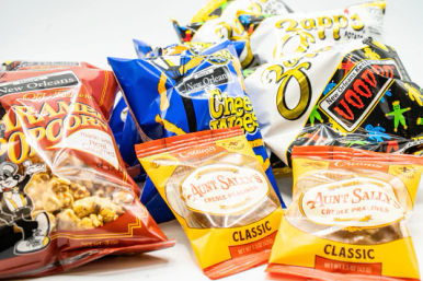 Colorful assortment of New Orleans–style snack bags on a white background: cheese puffs, potato chips, kettle popcorn and Creole praline candy.