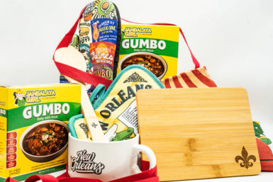 New Orleans–style gift set with gumbo mix boxes, a mug reading New Orleans holding a wooden spoon, colorful Cajun-themed oven mitts, a striped apron, and a small bamboo cutting board with a fleur-de-lis.