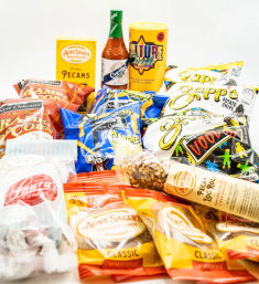 New Orleans‑style snack assortment on white background: colorful chip and cheese snack bags, pralines, candied pecans, a pecan log and hot sauce.