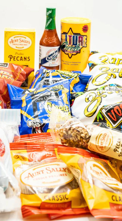 New Orleans‑style snack assortment on white background: colorful chip and cheese snack bags, pralines, candied pecans, a pecan log and hot sauce.