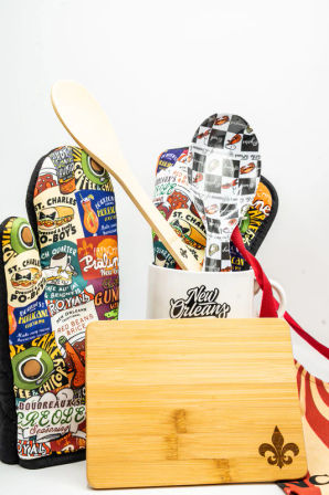 New Orleans–themed kitchen set with colorful printed oven mitts, a mug holding wooden and patterned serving spoons, and a bamboo cutting board engraved with a fleur-de-lis.