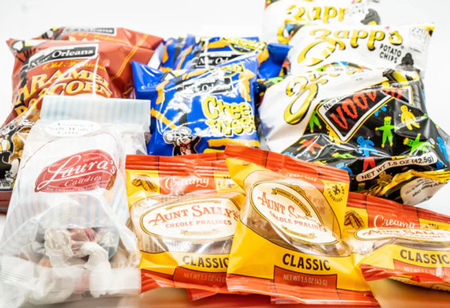 Assorted colorful New Orleans-style snack bags and individually wrapped Creole pralines on a white background, featuring potato chips, cheese snacks and classic candies.