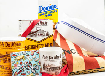 New Orleans–themed beignet and coffee gift set with beignet mix box, powdered sugar, yellow coffee tins, souvenir mug, colorful illustrated postcard, striped tote and paper boat hat on white background
