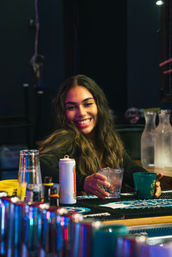 Happy woman at a neon-lit bar holding a glass of ice, smiling across a busy bartop with cups, bottles and cocktail tools — casual nightlife scene.