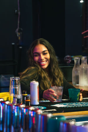 Happy woman at a neon-lit bar holding a glass of ice, smiling across a busy bartop with cups, bottles and cocktail tools — casual nightlife scene.