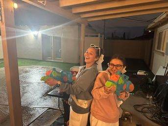 Two friends on a suburban backyard patio at night, laughing and aiming bright green-and-orange toy water guns under a covered porch with wet concrete