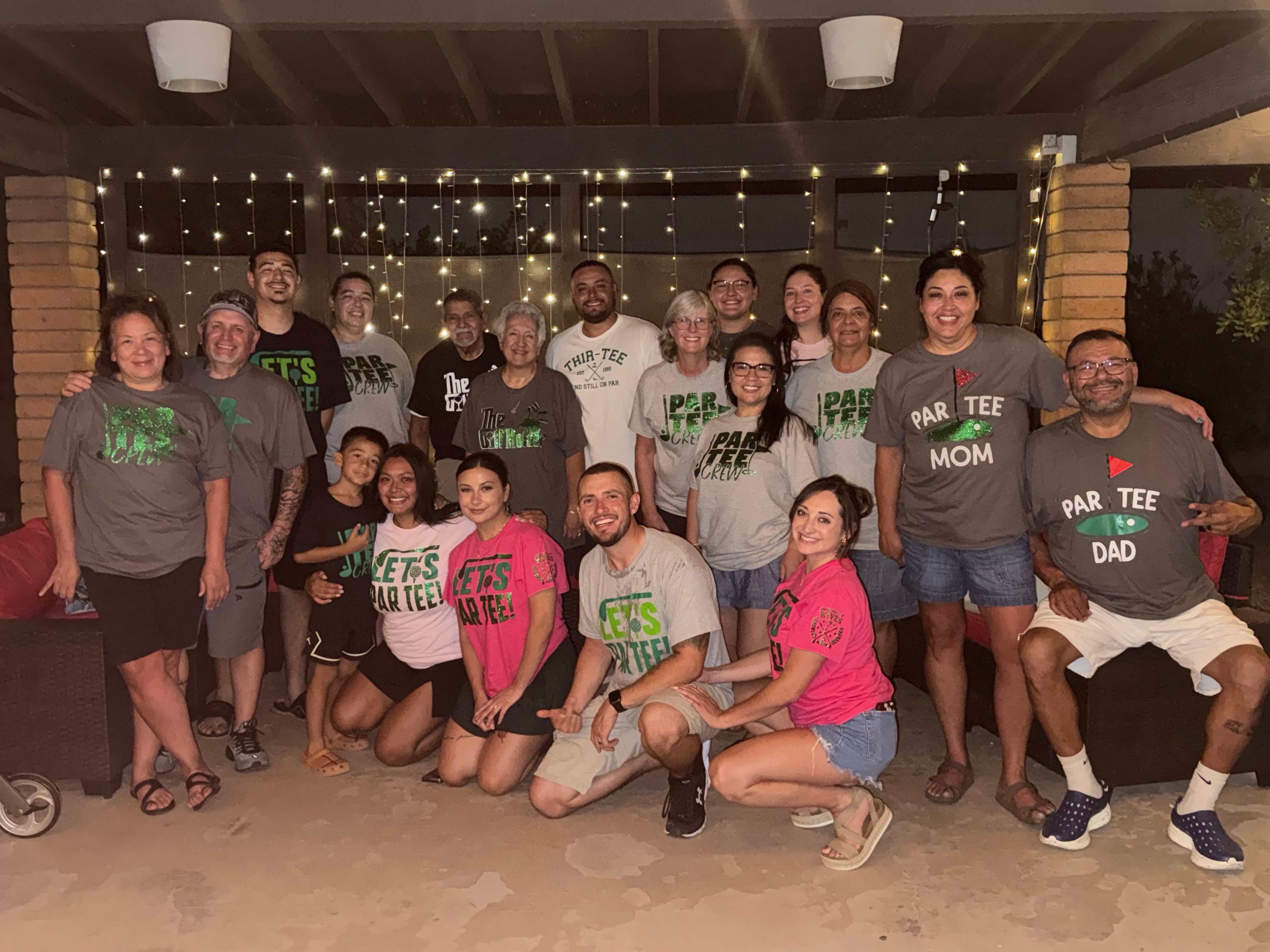 Cheerful multigenerational family reunion on a backyard patio at night, group smiling in matching 'Par Tee' themed shirts under warm string lights