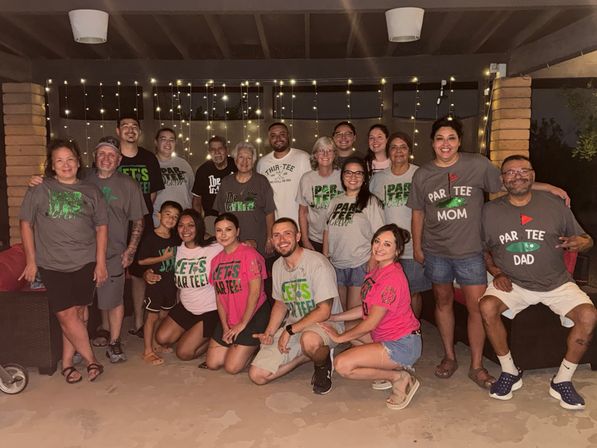 Cheerful multigenerational family reunion on a backyard patio at night, group smiling in matching 'Par Tee' themed shirts under warm string lights
