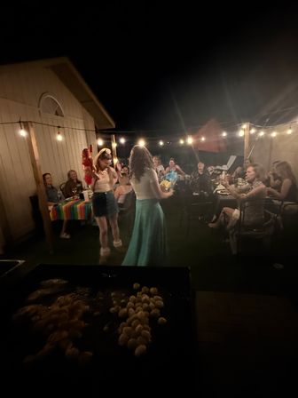 Cozy backyard patio party at night under string lights — friends seated at a long table while two people dance in the center, colorful tablecloth on one table and a griddle of sizzling snacks in the foreground.