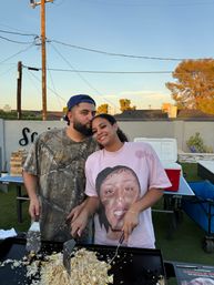 Smiling couple cooking fried rice on a large outdoor griddle in a suburban backyard at sunset — man kisses woman’s forehead while they flip food, cooler and ping-pong table behind them, power lines and trees overhead.