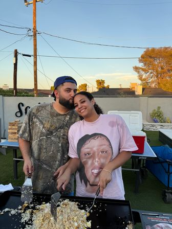 Smiling couple cooking fried rice on a large outdoor griddle in a suburban backyard at sunset — man kisses woman’s forehead while they flip food, cooler and ping-pong table behind them, power lines and trees overhead.