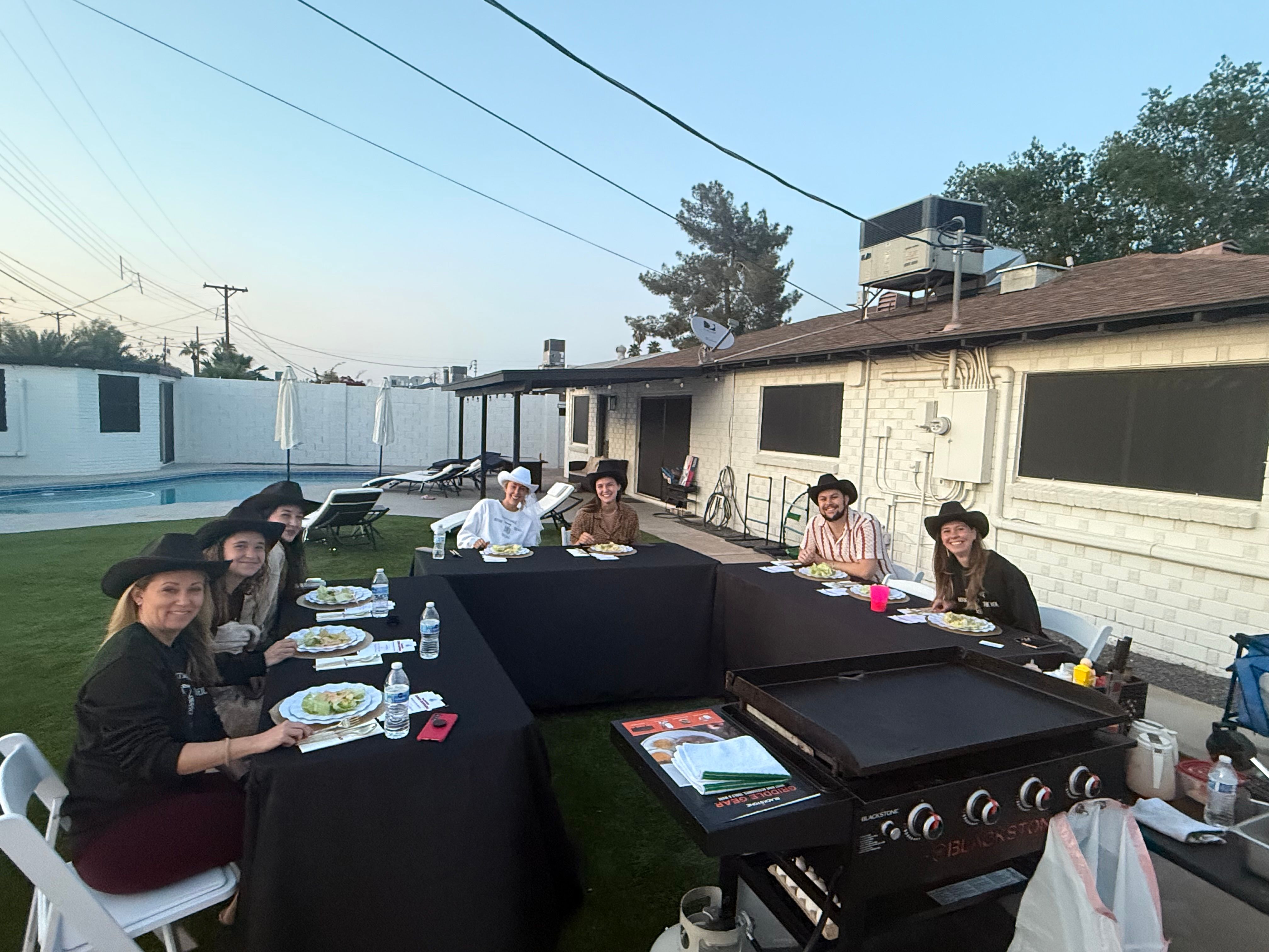 Backyard poolside barbecue at dusk: eight people wearing cowboy hats seated around U-shaped black tables with plates and water, large flat-top griddle in foreground and white brick house behind.