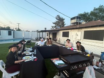 Backyard poolside barbecue at dusk: eight people wearing cowboy hats seated around U-shaped black tables with plates and water, large flat-top griddle in foreground and white brick house behind.